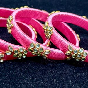 May include: A set of five vibrant pink bangles. Each bangle is wrapped in pink thread and adorned with gold-colored floral embellishments set with small, clear gemstones. The bangles are displayed on a dark blue surface.