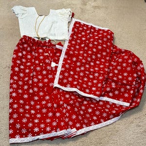 Puede incluir: Un conjunto de tres piezas con una parte superior blanca de manga corta con un patrón geométrico, una falda roja con diseños florales blancos y una bufanda roja a juego. La falda y la bufanda tienen ribetes de encaje blanco.