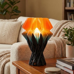 May include: A decorative table lamp with an orange and blue geometric shade and a black, textured base. The lamp is lit, casting a warm glow. It sits on a wooden table in a living room setting, next to books and a coaster.