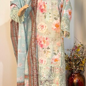 Può includere: Un completo composto da tunica a maniche lunghe e pantaloni azzurri con stampa floreale rosa e arancione. La tunica presenta un bordo in pizzo decorativo lungo lo scollo e l'orlo, abbinata a uno scialle coordinato con un bordo rosso scuro.