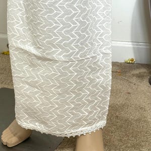 Puede incluir: Una falda larga blanca con un patrón geométrico y un delicado ribete de encaje en el dobladillo. La falda está hecha de una tela ligera, adecuada para climas cálidos. El diseño presenta un patrón repetitivo en forma de chevrón.