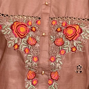 Op de afbeelding: Een bruin katoenen top met een bloemenborduursel. De borduursels bevatten roze, oranje en rode bloemen met groene bladeren. De top heeft een knoopsluiting.