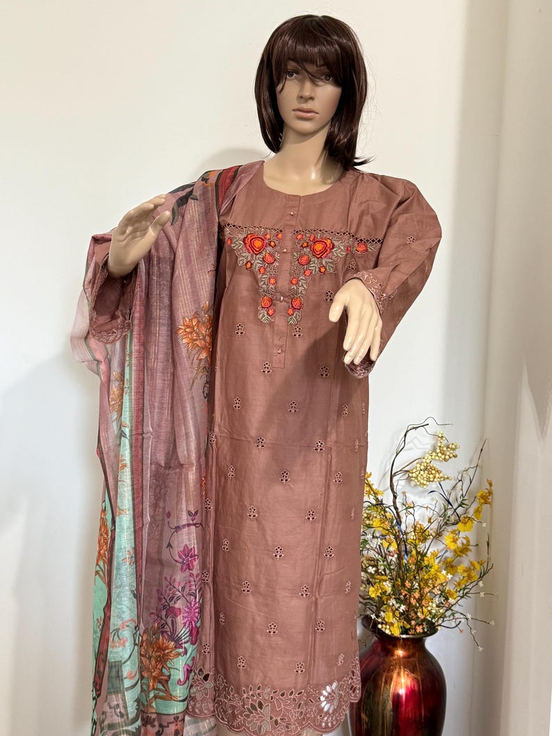 Op de afbeelding: Een bruine geborduurde jurk met een dupatta met bloemenprint. De jurk heeft een ronde halslijn en lange mouwen. De dupatta is lichtroze en groen met bloemenontwerpen.