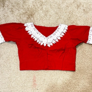 May include: A red blouse with a V-neckline and short sleeves. The neckline and sleeve edges are trimmed with white lace. The blouse is made of a smooth fabric and is laid flat on a neutral surface.
