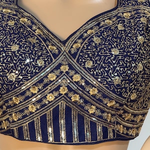Puede incluir: Top azul marino con intrincados bordados dorados y lentejuelas. El diseño presenta patrones florales y rayas verticales, creando un aspecto texturizado y ornamentado. El escote es cuadrado.