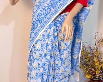 Può includere: Un sari azzurro e bianco con un motivo floreale è drappeggiato su un manichino. Il sari ha un bordo bianco ed è abbinato a una camicetta rossa. Un vaso marrone con fiori gialli è sullo sfondo.