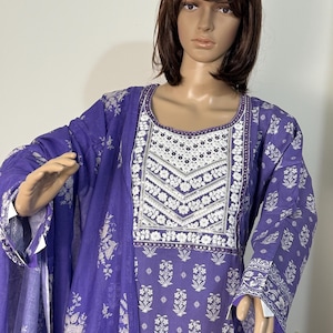 May include: A purple printed salwar kameez ensemble. The outfit features a long-sleeved tunic with a white floral design on the neckline and bodice. The matching dupatta and pants have a similar floral pattern. The fabric appears to be lightweight.