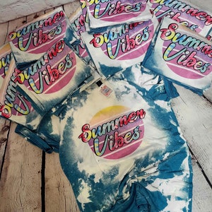 Puede incluir: Una pila de camisetas de color azul y blanco con efecto tie-dye con el texto "Summer Vibes" en una fuente retro y un gr&aacute;fico de puesta de sol arco&iacute;ris.