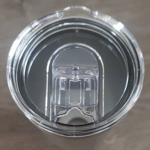 May include: Close-up of a clear plastic lid for a travel mug. The lid has a black rubber seal and a small hole in the center.