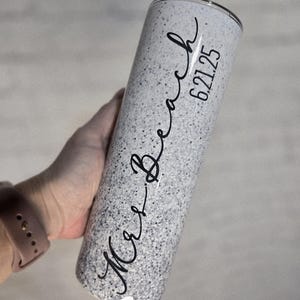 Puede incluir: Un vaso moteado blanco y gris con las palabras "Mrs. Beach" y la fecha "6.21.25" en escritura negra. El vaso de acero inoxidable es cilíndrico y parece estar aislado.