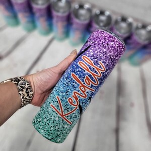 Puede incluir: Un vaso personalizado con purpurina degradada, con el nombre "Kendall" en letras naranjas. El vaso tiene un degradado de purpurina turquesa, azul, morado y rosa.