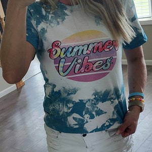 Puede incluir: Una camiseta azul turquesa tie-dye con el texto "Summer Vibes" en una fuente retro colorida. La camiseta tiene un fondo blanco con detalles tie-dye azules.