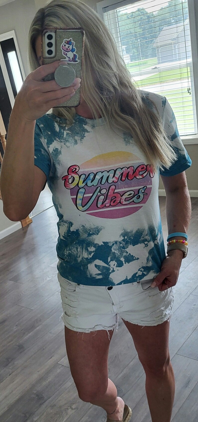 Puede incluir: Una camiseta azul turquesa tie-dye con el texto "Summer Vibes" en una fuente retro colorida. La camiseta tiene un fondo blanco con detalles tie-dye azules.