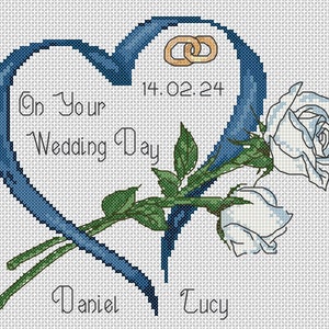 May include: A blue ribbon in the shape of a heart with two gold rings and the date 14.02.24. The ribbon is intertwined with two white roses and the names Daniel and Lucy. The text "On Your Wedding Day" is written inside the heart.