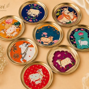 May include: A collection of eight circular brass-rimmed coasters featuring colorful illustrations of cows and calves amidst lotus flowers. Each coaster displays distinct artistic scenes with vibrant backgrounds in blue, orange, red, and magenta.