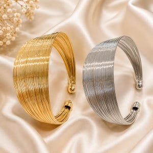 May include: Two wide cuff bracelets, one in a polished gold tone and one in a polished silver tone. Each bracelet is constructed from multiple thin, parallel wire strands that form a curved, open-ended shape with rounded ball-style end caps.