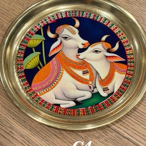 May include: A round brass plate featuring a central painting of two white cows against a dark blue background. The cows are adorned with orange and gold decorative blankets and necklaces. The border has a repeating pattern of red, green, and gold.