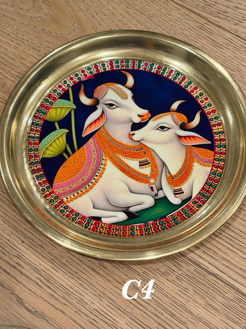 May include: A round brass plate featuring a central painting of two white cows against a dark blue background. The cows are adorned with orange and gold decorative blankets and necklaces. The border has a repeating pattern of red, green, and gold.