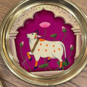 May include: A circular wall art piece featuring a gold frame and a central illustration of a white cow with orange handprints on its body. The cow stands against a vibrant magenta background with pink lotus flowers and an architectural archway design.