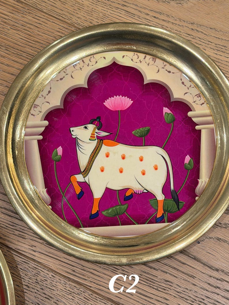 May include: A circular wall art piece featuring a gold frame and a central illustration of a white cow with orange handprints on its body. The cow stands against a vibrant magenta background with pink lotus flowers and an architectural archway design.