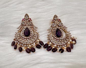 May include: A pair of elaborate gold-coloured earrings. Each earring has a large, teardrop-shaped, dark purple gemstone at the centre, encircled by smaller clear stones. Further dark purple beads and white pearls hang from the base.