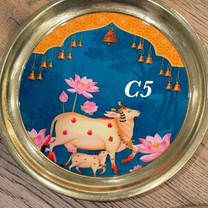 May include: A round brass tray featuring a central illustration of a cow and calf surrounded by pink lotus flowers. The background is a deep blue with an orange archway at the top adorned with hanging bells. The text C5 is printed in white.