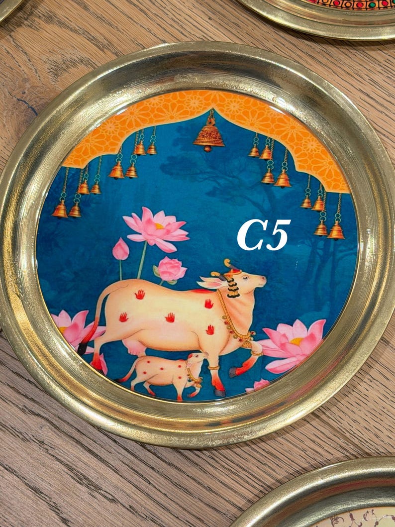 May include: A round brass tray featuring a central illustration of a cow and calf surrounded by pink lotus flowers. The background is a deep blue with an orange archway at the top adorned with hanging bells. The text C5 is printed in white.