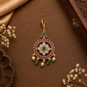 May include: A gold-toned pendant featuring a circular frame set with square-cut magenta stones. The center contains a floral arrangement of clear white stones and a central green stone. The base is adorned with dangling white pearls and green beads.