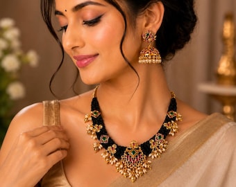 May include: A gold-toned necklace and matching jhumka earrings set featuring a thick black braided cord base. The jewellery is adorned with small green and red square-cut stones, clear crystals, and dangling white pearl-like beads in a cluster.