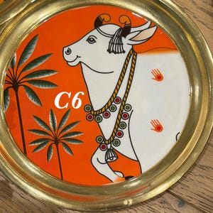 May include: A round brass tray featuring a vibrant orange background with a white cow illustration. The cow wears a beaded necklace and has small hand-shaped symbols on its side. Two palm trees are depicted on the left, and the text C6 is visible.