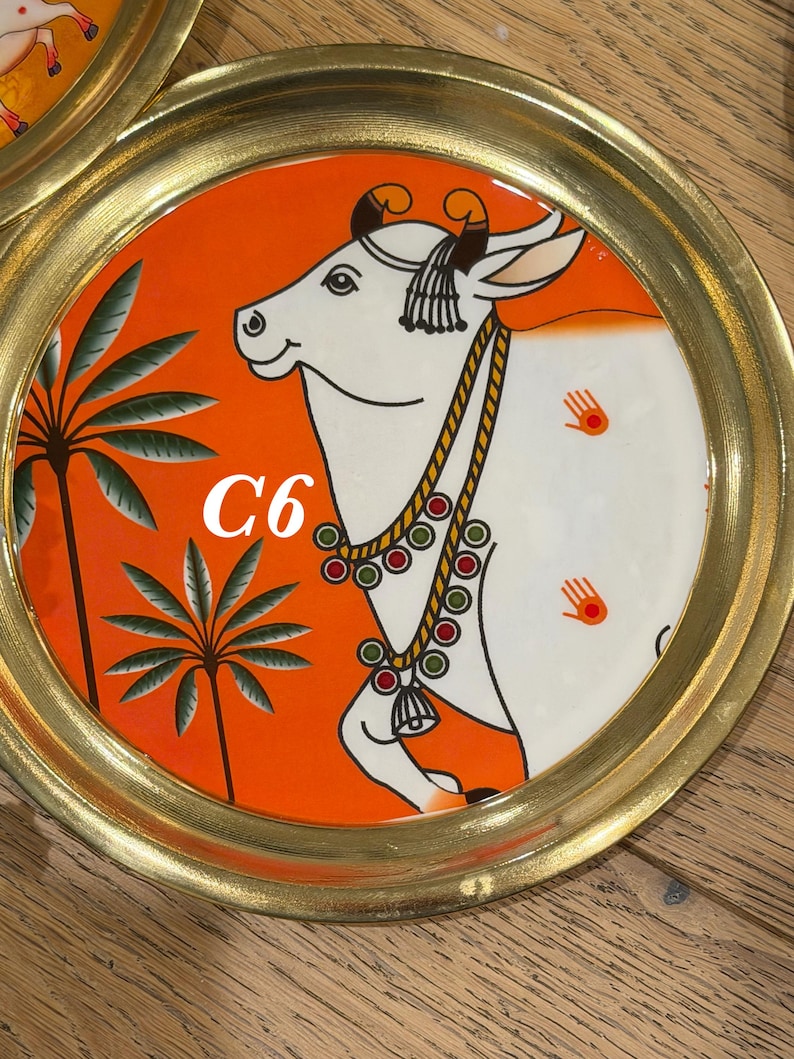 May include: A round brass tray featuring a vibrant orange background with a white cow illustration. The cow wears a beaded necklace and has small hand-shaped symbols on its side. Two palm trees are depicted on the left, and the text C6 is visible.