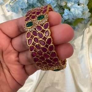 May include: A gold-toned bangle bracelet featuring a repeating pattern of teardrop-shaped deep pink stones. The design includes accents of small clear stones and two prominent oval-shaped green stones set within a decorative gold metal frame.