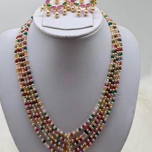 May include: A multi-strand necklace and matching earrings set. The necklace features strands of colorful beads and pearls, with a gold-toned pendant. The earrings have a gold-toned hoop design with pearls and colorful accents. The jewelry is displayed on a white bust.