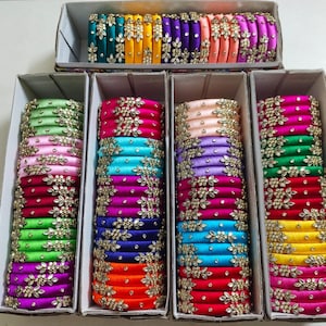 May include: A collection of silk-wrapped bangles arranged in five rectangular cardboard boxes. Each bangle features a smooth, vibrant fabric finish adorned with intricate, floral-shaped clear crystal embellishments in a repeating pattern.