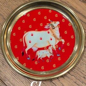 May include: A round brass tray featuring a vibrant red center inlay decorated with a repeating gold paisley pattern. The central illustration depicts a white cow and calf adorned with jewelry, accented by small red handprint motifs on the cow.