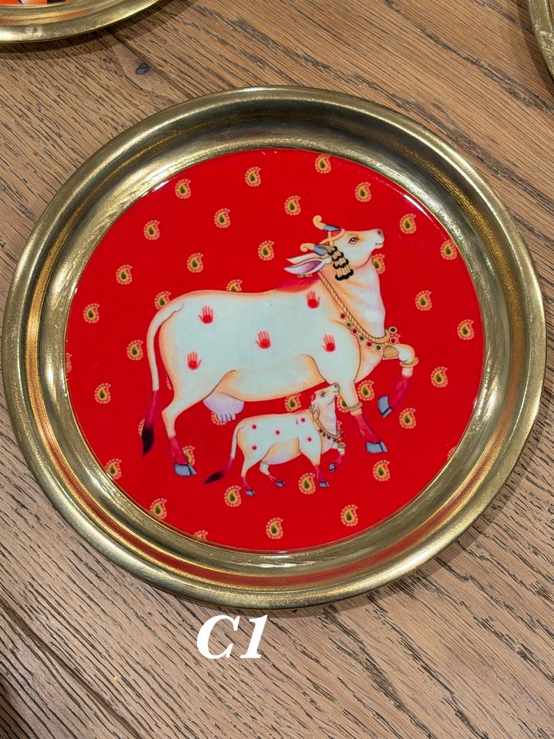 May include: A round brass tray featuring a vibrant red center inlay decorated with a repeating gold paisley pattern. The central illustration depicts a white cow and calf adorned with jewelry, accented by small red handprint motifs on the cow.
