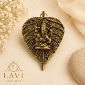 May include: A gold-toned metal pendant shaped like a textured leaf, featuring a central embossed figure of the deity Ganesha. The pendant has a small loop at the top for attachment and displays intricate vein patterns and a stippled surface finish.