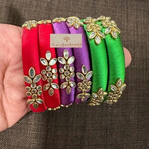 以下が含まれることがあります： 鮮やかな赤、紫、緑のシルク糸で巻かれた6つのバングルのセット。各バングルには、透明な石が付いた葉のクラスターのような形のゴールドトーンの金属装飾が施されています。Lavifashionsというテキストが書かれた長方形のラベルが見えます。