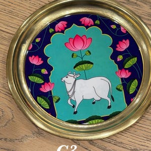 May include: A round decorative tray with a metallic gold rim and a central illustration featuring a white cow standing against a teal background. The design is framed by a dark blue border decorated with pink lotus flowers and green lily pads.