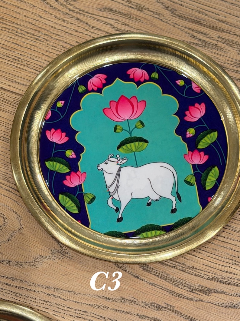 May include: A round decorative tray with a metallic gold rim and a central illustration featuring a white cow standing against a teal background. The design is framed by a dark blue border decorated with pink lotus flowers and green lily pads.