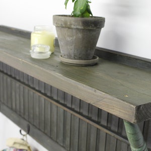 Op de afbeelding: Een rustiek houten wandrek met een groene geverfde afwerking. Het rek heeft een latten achterkant en haken aan de onderkant. Een kleine plant in een pot, een kaars en een glazen pot staan op het rek.