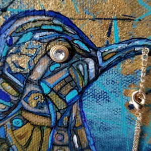 May include: Close-up of a mixed media painting featuring a bird's head. The artwork uses shades of blue, gold, and teal. A small, jeweled button serves as the bird's eye. A silver chain hangs on the right side of the painting.