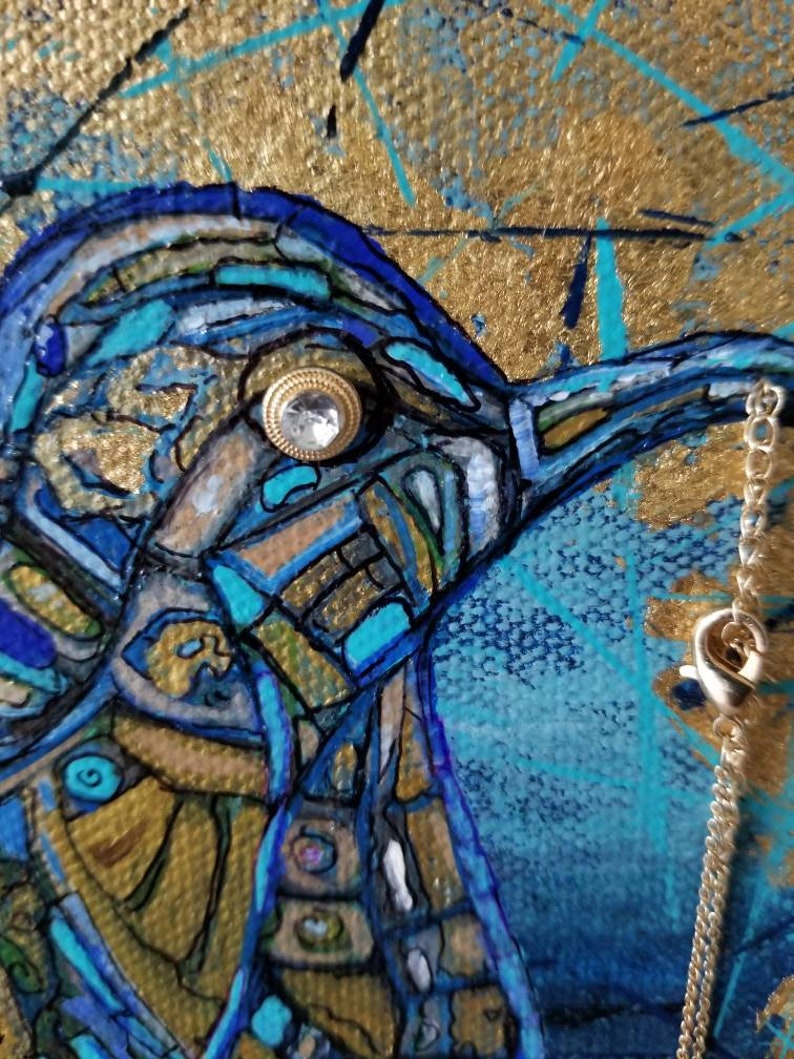May include: Close-up of a mixed media painting featuring a bird's head. The artwork uses shades of blue, gold, and teal. A small, jeweled button serves as the bird's eye. A silver chain hangs on the right side of the painting.