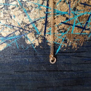 May include: A gold-colored necklace with a double-circle pendant, encrusted with small, clear gemstones. The necklace hangs against a backdrop of a dark blue canvas with gold and turquoise paint strokes.