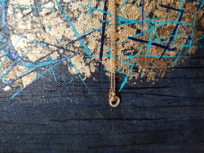 May include: A gold-colored necklace with a double-circle pendant, encrusted with small, clear gemstones. The necklace hangs against a backdrop of a dark blue canvas with gold and turquoise paint strokes.