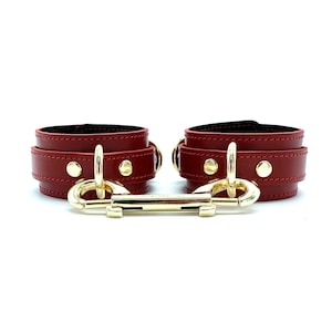 May include: A pair of red leather wrist cuffs with gold metal accents and a connecting clasp.