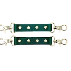 May include: Two dark green leather straps with silver-colored swivel clasps and rivets. The straps are designed to connect to a bag or other accessory. The background is plain white.