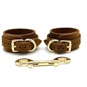 Puede incluir: Un par de puños de cuero marrón con hebillas doradas y una cadena de conexión de metal dorado.