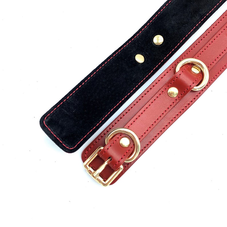 May include: A red leather collar with gold hardware and a black suede lining. The collar has two rings and a buckle.