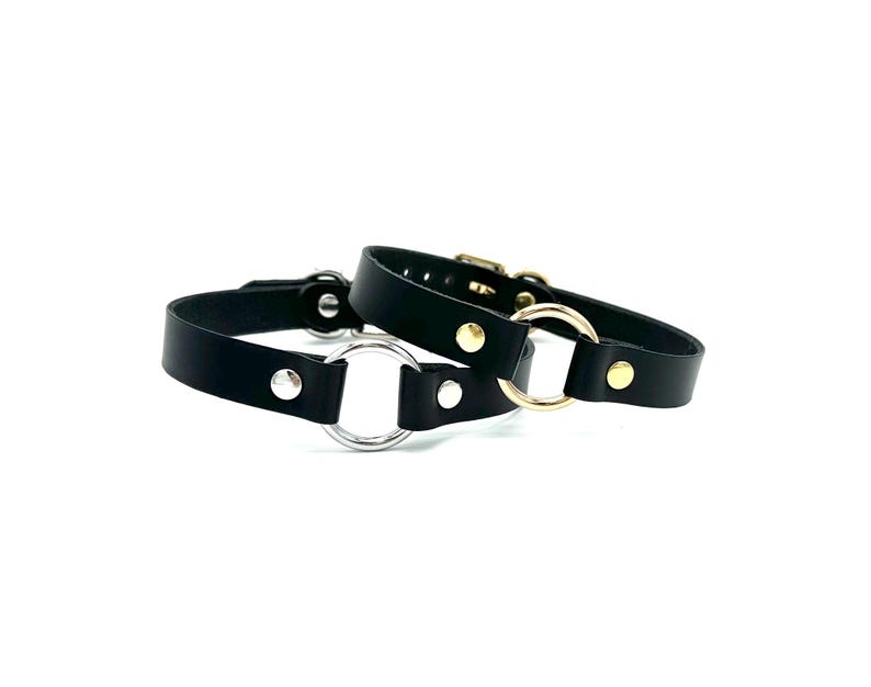 May include: Two black leather collars with metal rings. One collar has a silver ring and the other has a gold ring.